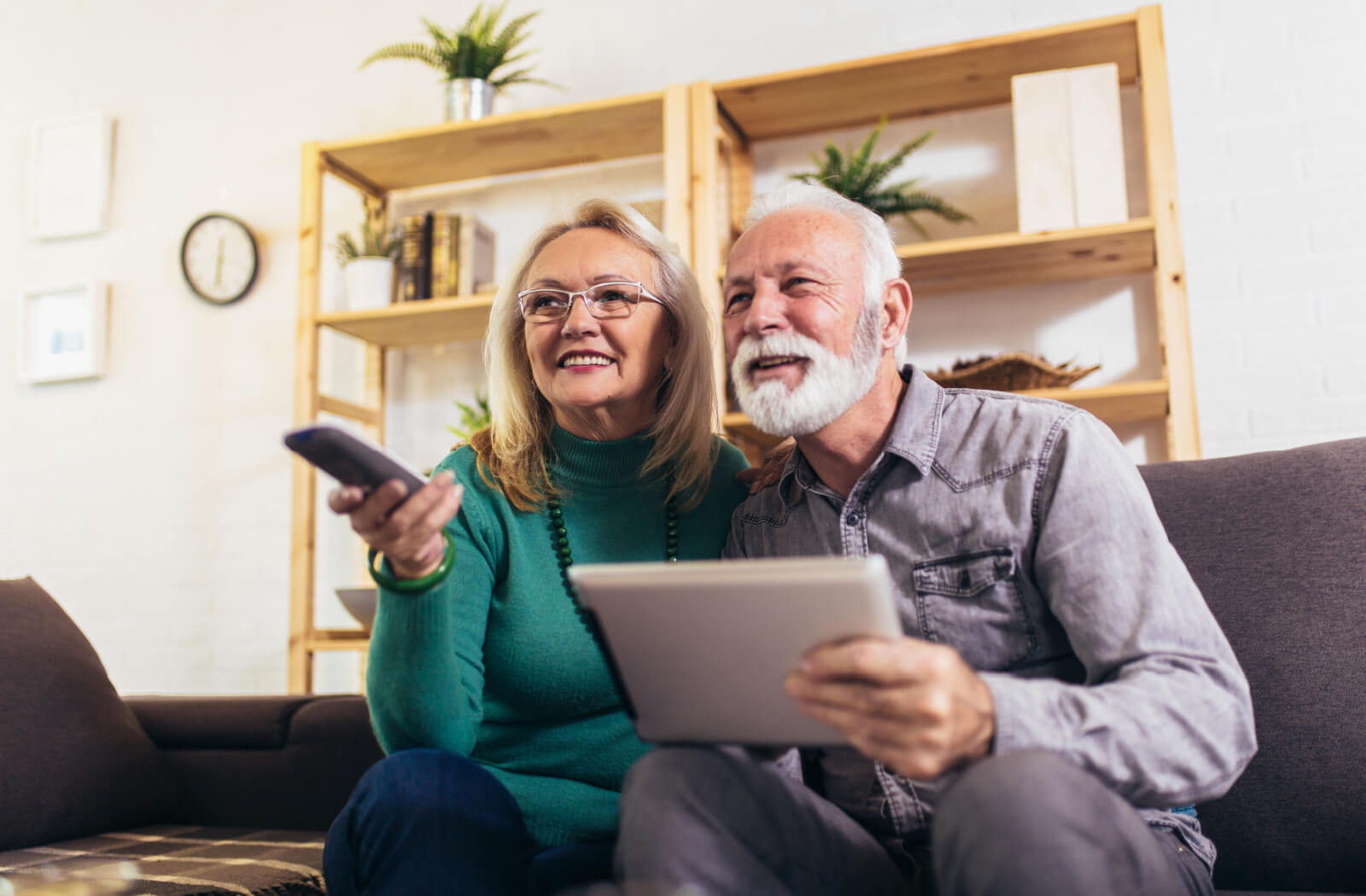 A senior couple smiling, holding a remote and a tablet, getting ready to watch a movie about Alzheimer's disease.