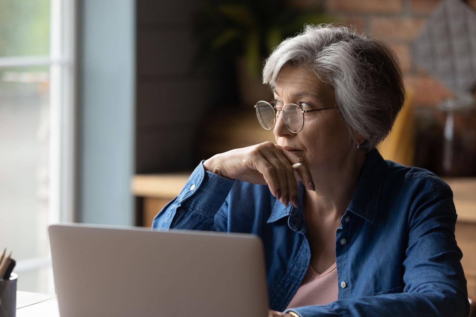 A senior lady thoughtfully looking away from her laptop out the window.