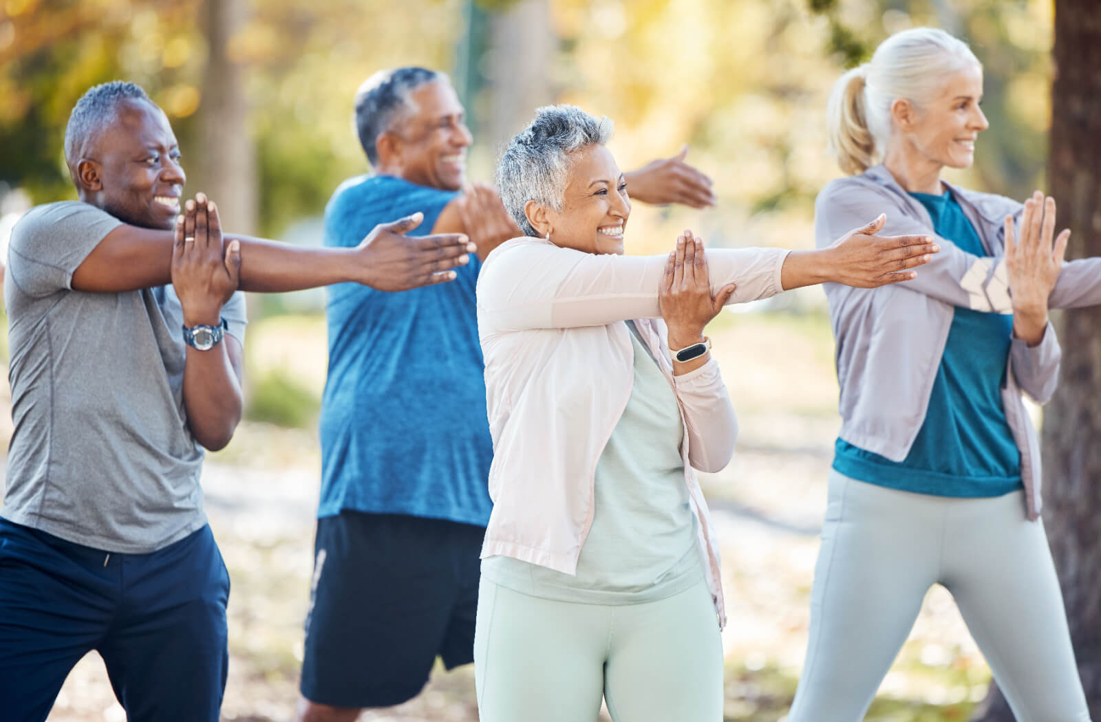 A group of 4 older adults smiling in the park, stretching their arms and getting ready for a low-impact workout together.
