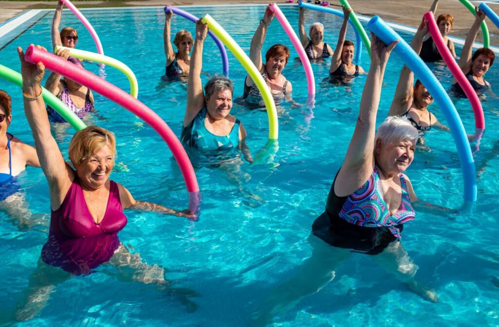 A group of older adult women exercising in a pool using swimming noodles.