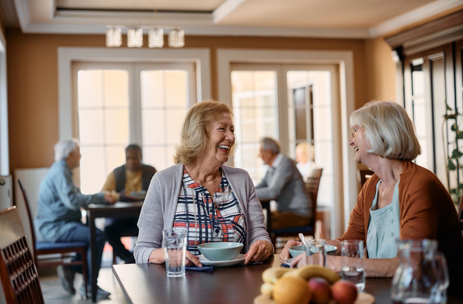 2 older adults smiling while chatting over breakfast in a common area in an assisted living community.