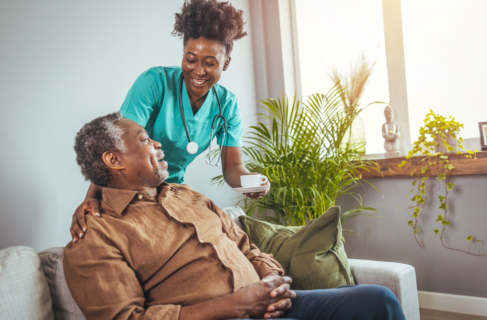 An older adult sitting on a couch while a caregiver in blue scrubs smiles and hands them a cup of coffee.