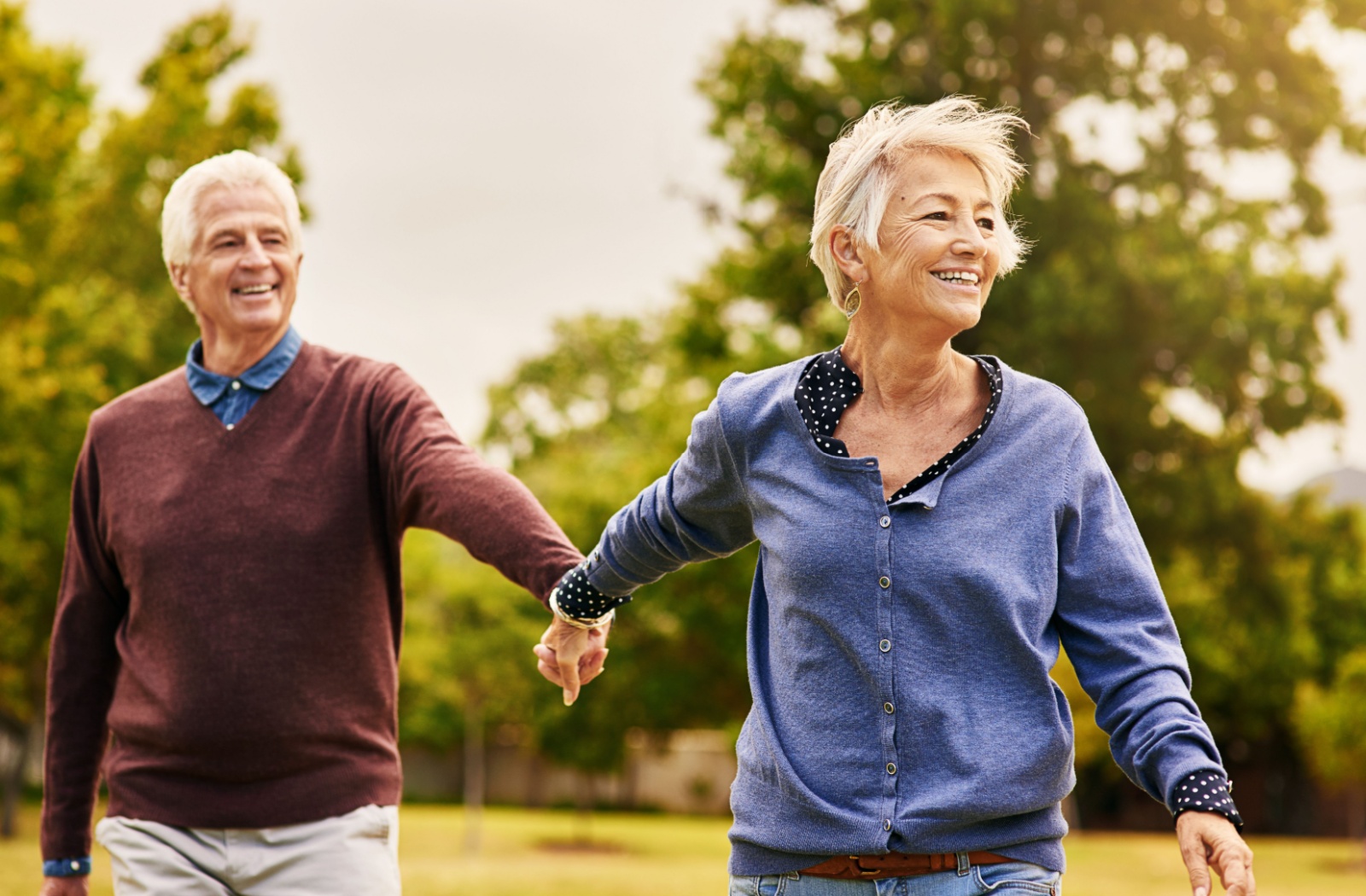 An older couple holding hands in the park and smiling while going on a summertime walk.