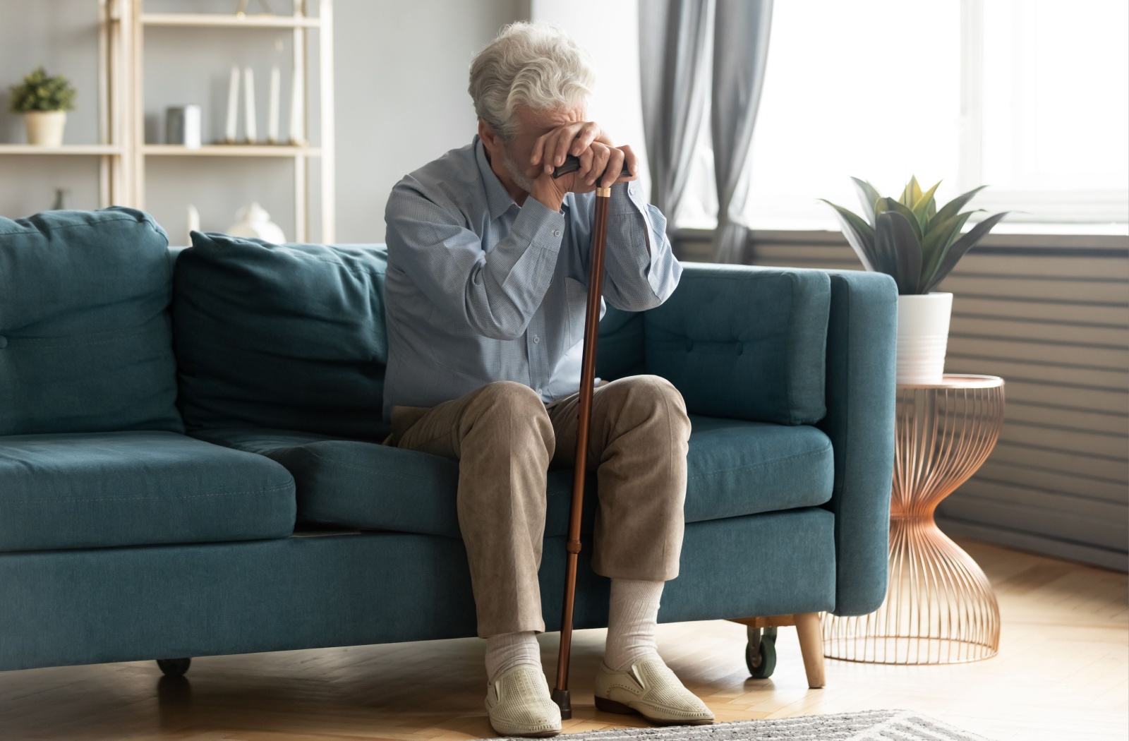 A senior sitting on a couch rests their head against the handle of their cane, frustrated at their growing walking difficulty