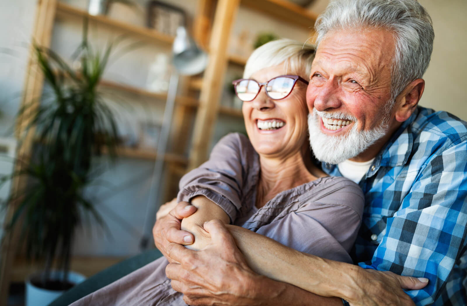 An older adult hugs their laughing spouse from behind in their well-decorated living room in front of a shelf of plants and picture frames
