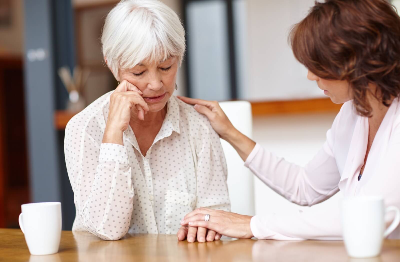 An older woman at a table looking down and her adult daughter resting her hand on her mother's shoulder while looking at her