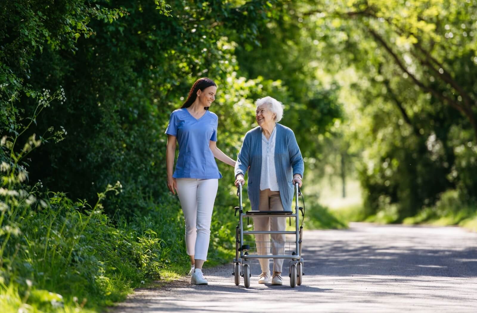 An older adult with a walker takes a stroll along a garden path with a smiling caregiver outside of a memory care community
