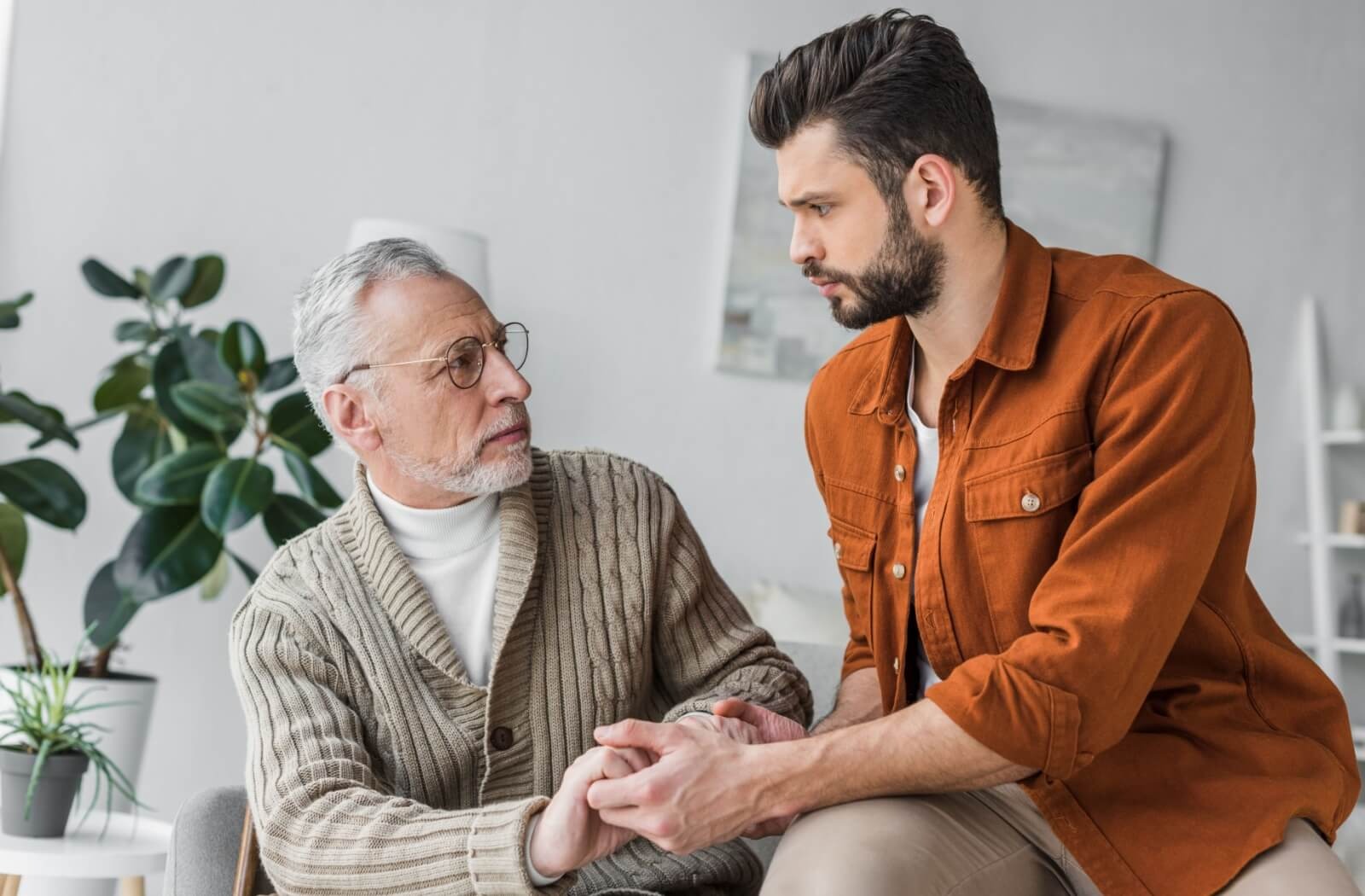 An adult child clasps their older parents hands while sitting beside one another during a conversation about caregiver burnout
